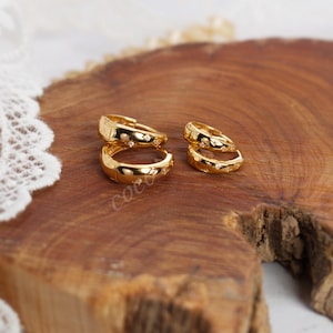 May include: Two pairs of gold hoop earrings with a polished finish and small, embedded star-shaped accents. Each hoop features a hinged closure mechanism. The earrings are arranged in two sets of different sizes, showcasing a smooth, reflective surface.