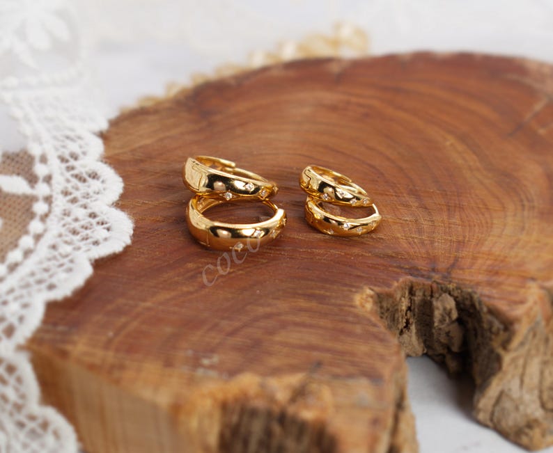 May include: Two pairs of gold hoop earrings with a polished finish and small, embedded star-shaped accents. Each hoop features a hinged closure mechanism. The earrings are arranged in two sets of different sizes, showcasing a smooth, reflective surface.