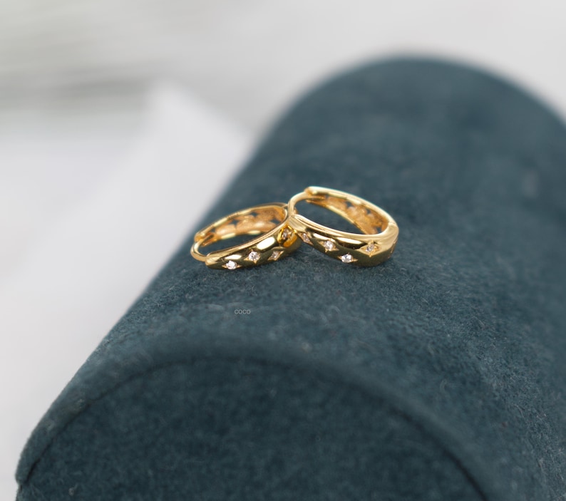 May include: A pair of gold hoop earrings featuring a polished finish and small, star-shaped cutouts. Each star cutout contains a tiny, clear, sparkling gemstone set into the centre of the gold hoop, creating a subtle and reflective design.