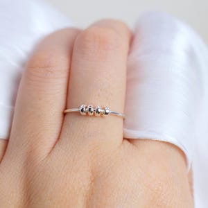 May include: A silver ring with a thin band and four small, rounded beads. The ring is shown on a finger, with a white fabric in the background. The beads are evenly spaced on the ring.