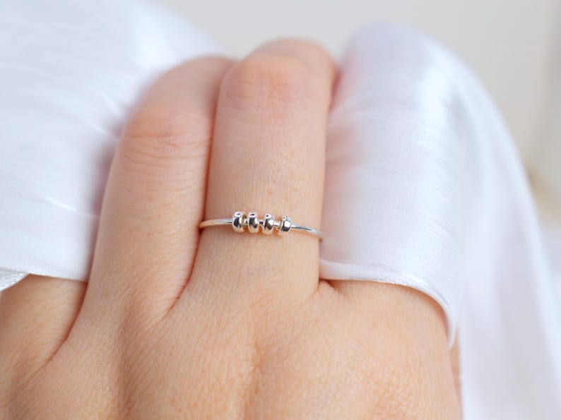 May include: A silver ring with a thin band and four small, rounded beads. The ring is shown on a finger, with a white fabric in the background. The beads are evenly spaced on the ring.