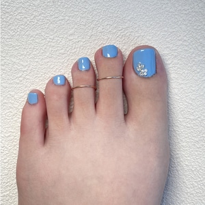 May include: A foot with light blue nail polish on all five toenails. The big toenail features a decorative cluster of small clear crystals and white pearls. Two thin, plain metallic toe rings are worn on the second and third toes.