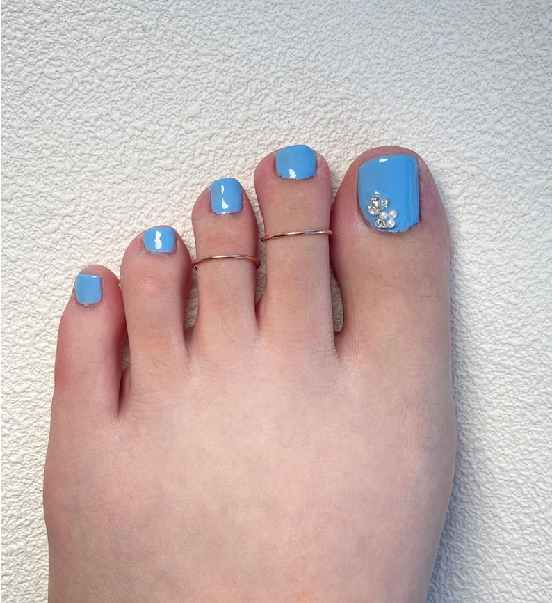 May include: A foot with light blue nail polish on all five toenails. The big toenail features a decorative cluster of small clear crystals and white pearls. Two thin, plain metallic toe rings are worn on the second and third toes.