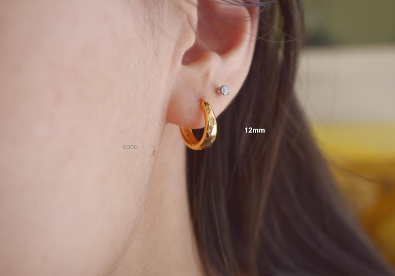 May include: A gold hoop earring with a 12mm diameter, featuring a series of small, embedded clear stones arranged in a star-like pattern along the outer surface. The earring is worn in the earlobe, accompanied by a small stud earring above it.