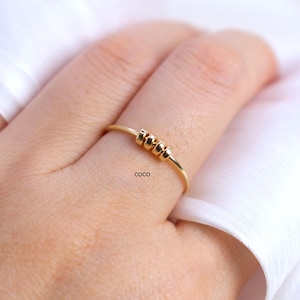 May include: A delicate gold ring with three small, textured beads centered on the band. The ring is worn on a finger, with the word "coco" printed on the finger. The background is a soft, white fabric.