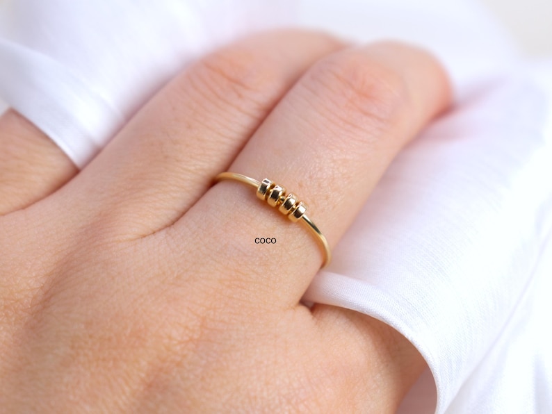 May include: A delicate gold ring with three small, textured beads centered on the band. The ring is worn on a finger, with the word "coco" printed on the finger. The background is a soft, white fabric.