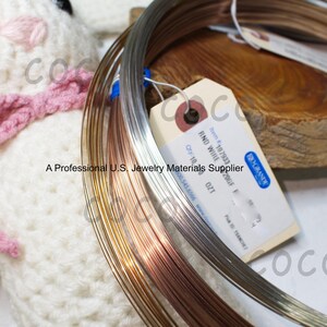 May include: Coils of round wire in silver, gold, and copper tones are displayed. A tag reads "RND WIRE" and "A Professional U.S. Jewelry Materials Supplier". A white knitted item with a pink bow is in the background.
