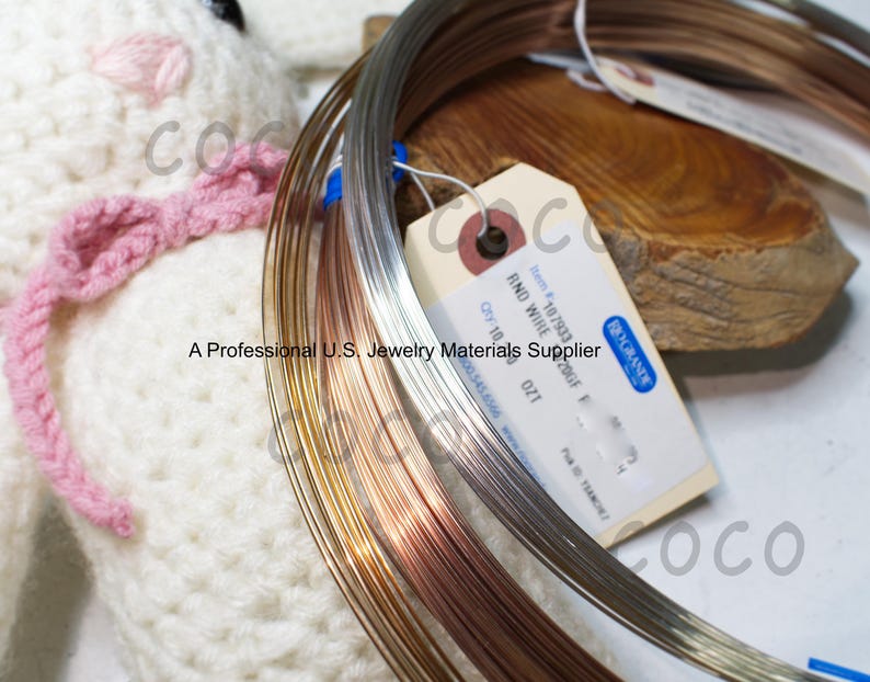 May include: Coils of round wire in silver, gold, and copper tones are displayed. A tag reads "RND WIRE" and "A Professional U.S. Jewelry Materials Supplier". A white knitted item with a pink bow is in the background.