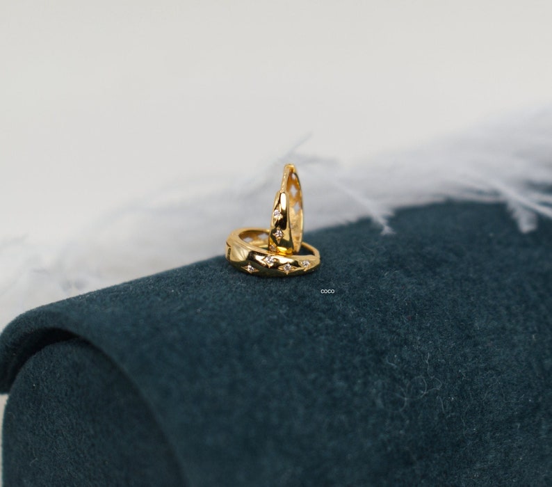 May include: A pair of gold hoop earrings featuring a polished finish and small, star-shaped engravings, each centred with a tiny clear gemstone. The earrings are arranged with one standing upright and the other resting horizontally.