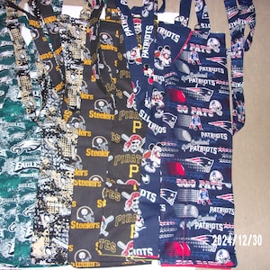 May include: A collection of seven fabric aprons with various sports team logos, including the Philadelphia Eagles, Pittsburgh Steelers, and New England Patriots. The aprons are made of cotton fabric and have adjustable straps.