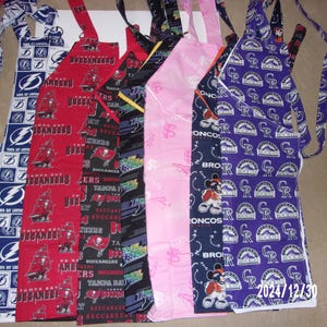 May include: Six different fabric aprons with various sports team logos and designs. The aprons are in blue, red, black, pink, purple, and white.