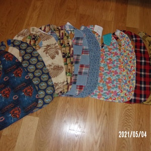 May include: A collection of 10 colorful fabric bibs with various patterns, including a Chicago Bears logo, wine bottles, plaid, and floral designs.