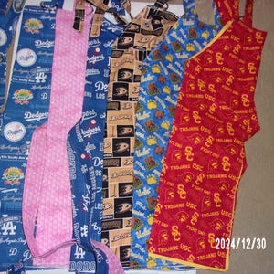 May include: Five fabric aprons with different designs. The aprons are made of cotton fabric and have adjustable straps. The designs include a pink floral pattern, a blue and white Dodgers pattern, a blue and white UCLA pattern, a brown and white duck pattern, and a red and yellow USC Trojans pattern.