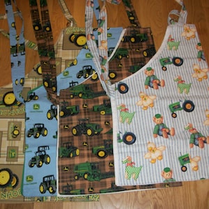 May include: Five different fabric aprons with various designs. Two aprons feature a brown and green plaid pattern with a John Deere tractor design. One apron has a blue and white striped pattern with a teddy bear and tractor design. Two aprons have a blue and white striped pattern with a teddy bear and tractor design.