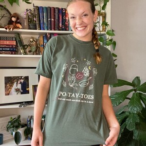 May include: A green t-shirt with a graphic of a cartoon character holding a frying pan and potatoes. The text "PO-TAY-TOES" is printed on the shirt, along with the phrase "boil em-mash em-stick em in a stew".