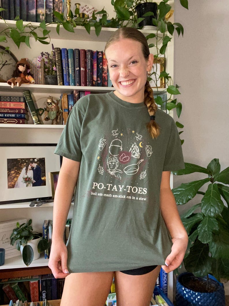 May include: A green t-shirt with a graphic of a cartoon character holding a frying pan and potatoes. The text "PO-TAY-TOES" is printed on the shirt, along with the phrase "boil em-mash em-stick em in a stew".
