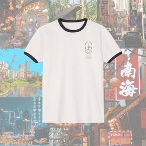 May include: White ringer t-shirt with black trim around the collar and sleeves. The shirt features a small graphic on the chest with the text "Twice Lucky Japanese Cuisine". The shirt is set against a background of Asian cityscapes.
