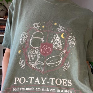 May include: A green t-shirt with a white graphic of a crest featuring a hobbit, potatoes, a frying pan, and a fire. The text "PO-TAY-TOES" is below the crest with the text "boil em-mash em-stick em in a stew" below that.