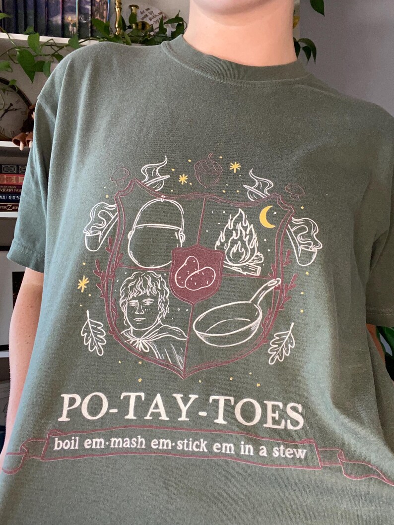 May include: A green t-shirt with a white graphic of a crest featuring a hobbit, potatoes, a frying pan, and a fire. The text "PO-TAY-TOES" is below the crest with the text "boil em-mash em-stick em in a stew" below that.
