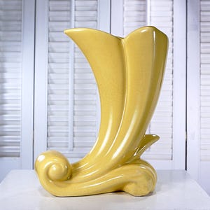 May include: A yellow ceramic vase with a unique, curved design. The vase features a flared top and a base with a swirling, wave-like detail. The vase is approximately 12 inches tall.