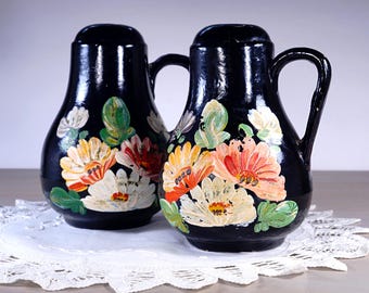 May include: Two black ceramic pitchers with rounded bodies and handles, each featuring hand-painted floral designs in shades of white, yellow, orange, and green. The pitchers have a glossy finish and are positioned side by side on a surface.