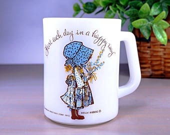 May include: White ceramic mug featuring a Holly Hobbie illustration of a girl in a blue bonnet holding flowers. The mug has the text "Start each day in a happy way." American Greetings Corp. 1972.