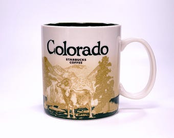 May include: White ceramic mug with a green rim and handle. The mug features a brown and gold illustration of a mountain landscape with a bighorn sheep in the foreground. The text "Colorado" is printed in black at the top of the mug, and "Starbucks Coffee" is printed in black below the landscape.