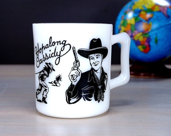 May include: White ceramic mug with a black and white illustration of Hopalong Cassidy, a cowboy with a gun, and another cowboy riding a horse. The mug has a black handle.