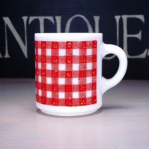 May include: White ceramic mug with a red and white gingham pattern. The mug has a handle and is sitting on a white surface.