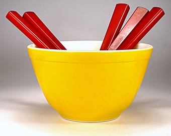May include: A yellow mixing bowl with a white rim, filled with red mixing spoons.