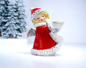 May include: A ceramic Christmas angel figurine with a red coat, white fur trim, and a red hat. The angel has blonde hair and is holding a white object. The figurine has small white wings and is set against a snowy background.