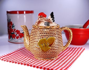 May include: A decorative teapot with a chicken-shaped lid, featuring a woven basket design. The teapot has a gold leaf-shaped accent with the word "TEA". It sits on a red and white checkered napkin, with a red bowl and a floral tin in the background.