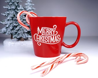 May include: A red ceramic mug with the words "MERRY CHRISTMAS" in white script. Two candy canes are in the mug and two are lying in front of it. A small, snow-covered Christmas tree is in the background.
