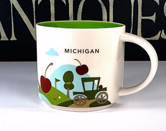 May include: White ceramic coffee mug with a green rim and a cartoon illustration of a car driving through a green field with red cherries and the word "MICHIGAN" on the front.