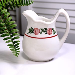 May include: White ceramic creamer with a red and green floral design. The creamer has a rounded body and a handle.