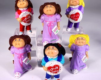 May include: Set of six Cabbage Patch Kids figurines. Each figurine is wearing a different outfit and holding a different accessory. The figurines are all in various shades of purple, pink, and blue.
