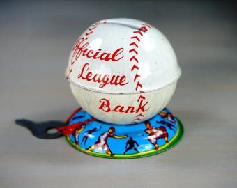May include: A white baseball-shaped coin bank with red stitching and the words "Official League Bank" printed on it. The bank has a keyhole and a base with a blue background and a scene of baseball players.