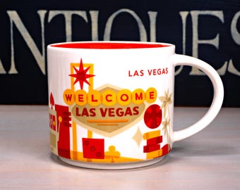 May include: White ceramic mug with a red interior and a curved handle. The mug features a colorful design with the words "WELCOME LAS VEGAS" and various iconic symbols in red, yellow, and orange. The words "LAS VEGAS" are also printed on the mug.