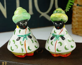 May include: A pair of vintage salt and pepper shakers in the shape of people wearing green and yellow hats and white and green patterned robes. The figures have black faces and are standing on a brown surface.