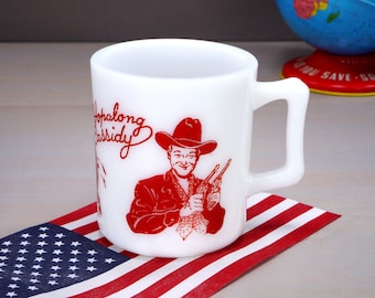 May include: White ceramic mug with a red illustration of a cowboy with a gun and the text "Hopalong Cassidy". The mug is sitting on a red, white, and blue American flag.