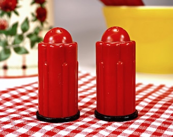 May include: Two red salt and pepper shakers with black tops, sitting on a red and white checkered tablecloth.