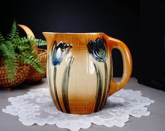 May include: A ceramic pitcher with a tulip design. The pitcher has a tan background with a brick-like texture and a curved handle. Blue and white tulips are painted on the front. The pitcher sits on a white lace doily.