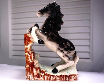 May include: A ceramic figurine of a black and white horse rearing up on its hind legs, with its front legs raised. The horse is standing on a brown base with a green and white painted design.