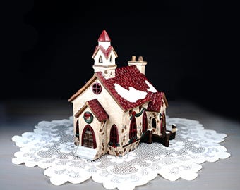 May include: A ceramic Christmas church figurine with a red roof, white snow accents, and arched windows. The church has a steeple and is decorated with a wreath and garland. The figurine is placed on a white lace doily.