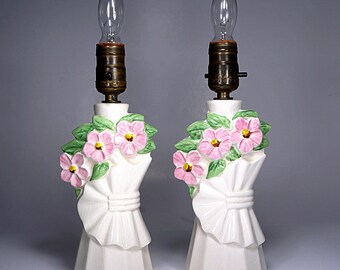 May include: Pair of vintage white ceramic table lamps. Each lamp features a floral bouquet design with pink flowers and green leaves. The lamps have a bow detail and a faceted base. The bulbs are visible.