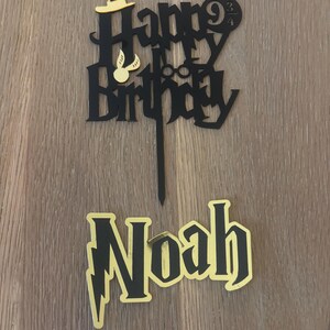 May include: A black and gold cake topper with the words "Happy Birthday" and "Noah". The topper features a wizard hat, glasses, and a lightning bolt. The cake topper is designed for a birthday celebration.