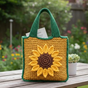 Può includere: Borsa tote all'uncinetto con un motivo a girasole. La borsa è gialla con un centro marrone e petali gialli. Ha manici e bordi verdi. Un libro è visibile all'interno della borsa. La borsa è su un tavolo di legno.