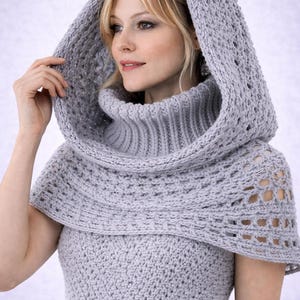 Può includere: Un cappuccio e un collo alto lavorati all'uncinetto grigio chiaro. Il cappuccio presenta un motivo strutturato e dettagli traforati. Il collo è indossato sopra un top all'uncinetto abbinato, creando un look coerente ed elegante.