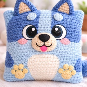 Puede incluir: Un cojín de crochet hecho a mano con forma de cara de perro. Presenta tonos azules, con detalles beige en las orejas y las patas. El perro tiene ojos grandes, mejillas rosas y una lengua rosa, creando una apariencia amigable.