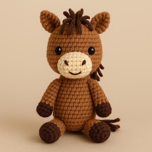 May include: A handmade crochet horse doll in shades of brown and tan. The horse has a light tan muzzle, dark brown hooves, and a dark brown mane and tail. The doll has black safety eyes and is sitting upright.